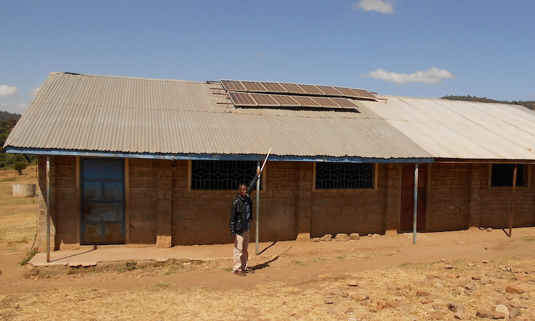 A Yiaku man points to solar panels atop a school where children are taught Yiaku language and culture. The Yiaku are working to preserve their traditional knowledge, which includes successful approaches to conserving Mukogodo Forest. to Image by Shadrack Kavilu for Mongabay.