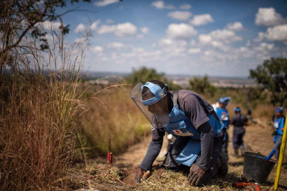 clearing mines in Angola, a HALO Trust worker wearing blue protective gear on his knees probing grass and dirt with his hands. Credit: The HALO Trust