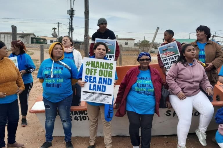 Protesting fisherfolk standing by a beached boat with a sign saying "Hands off our sea and land". Image courtesy Wendy Pekeur.