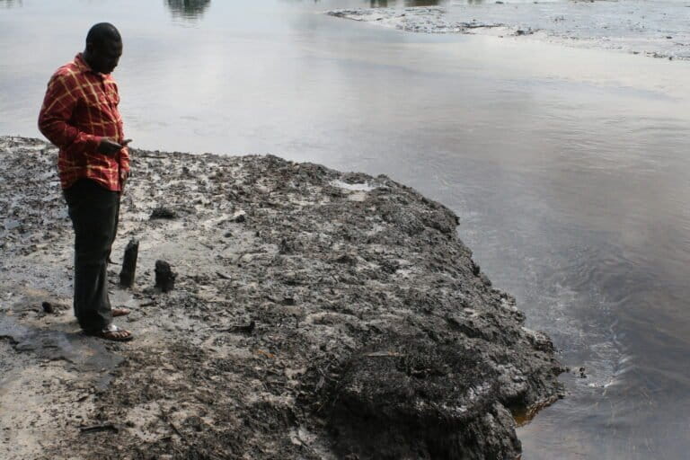 A man in a red shirt standing on barren grey brown shore, oily water of Goi Creek flowing past. Friends of the Earth International via Flickr (CC BY-NC-ND)