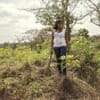 Dorothee Mbogo, carrying a machete and wearing a sleeveless white top and dark blue jeans, standing in the disturbed earth of her cassava farm in Batchenga, Cameroon. Image by Ryan Brown/UNWomen via Flickr (CC BY-NC-ND 2.0)