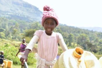 A young girl in a pale pink dress, a bobble hat and smile, pushing a wheelbarrow with two large yellow water containers across a field at Kapsambu village, Mt Elgon, Kenya. Image courtesy Infonile.