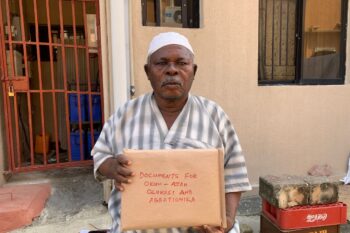 A stocky man in a white cap and grey and white dashiki holds up a thick manila envelope of documents of his demolished property, red marker announcing its contents. Image courtesy Kemi Adelagun.