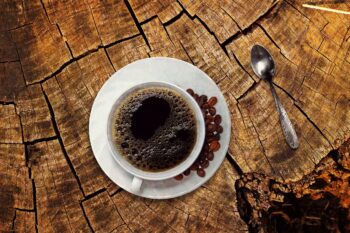 A cup of coffee with beans and a teaspoon on a stump tabletop. Image by Anja (cocoparisiene) from Pixabay (Pixabay Content License).
