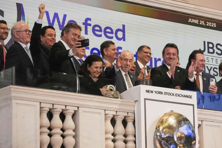 JBS founder José Batista Sobrinho (center) flanked by his sons, Joesley (taking a selfie) and Wesley (clapping), waves from the podium after ringing the New York Stock Exchange opening bell, Wednesday, June 25, 2025. Image by AP Photo/Richard Drew.