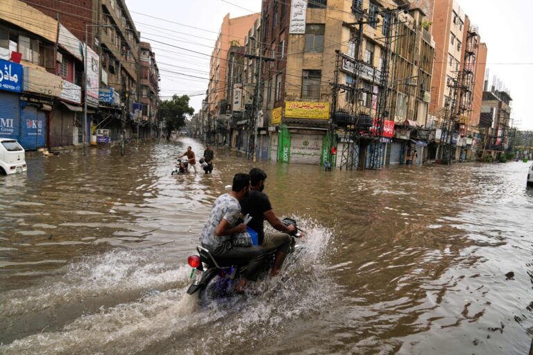 Banner image of motorists riding through a flooded road caused by heavy monsoon rain in Lahore, Pakistan, on July 10, 2025. Image by AP Photo/K.M. Chaudary.