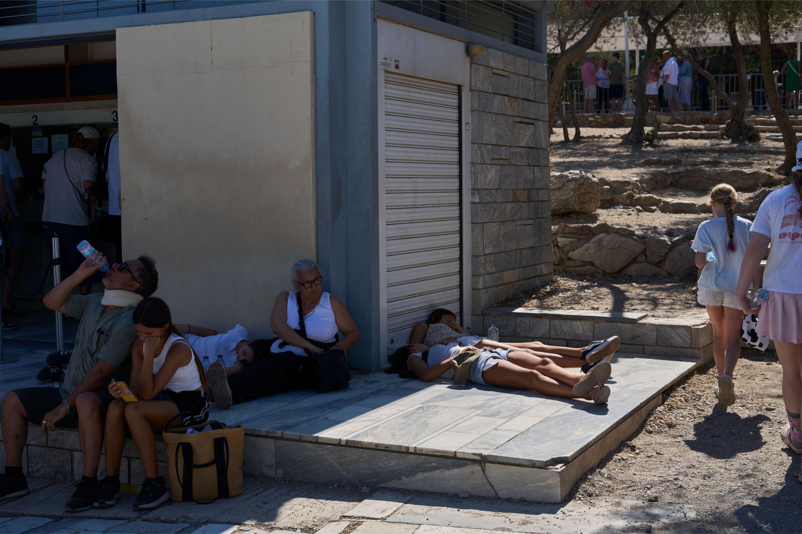 A tourist drinks water as others sleep outside the entrance of the Acropolis hill during a heat wave in Athens, July 9, 2025. Image by AP Photo/Petros Giannakouris