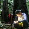 Jorian Hendricks in the forest of Brito Ulu, Indonesian Borneo, taking field notes in his notebook while using a directional microphone to record the sounds of the forest.