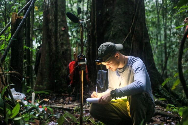 Jorian Hendricks in the forest of Brito Ulu, Indonesian Borneo, taking field notes in his notebook while using a directional microphone to record the sounds of the forest.