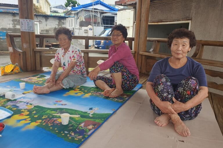 Haenyeo and best friends, from left, Lee Yeon-oak, Im Gil-seok, and Kim Choon-hee, in a seashore gazebo in Guman-ri, South Korea, on Aug. 16, 2026.