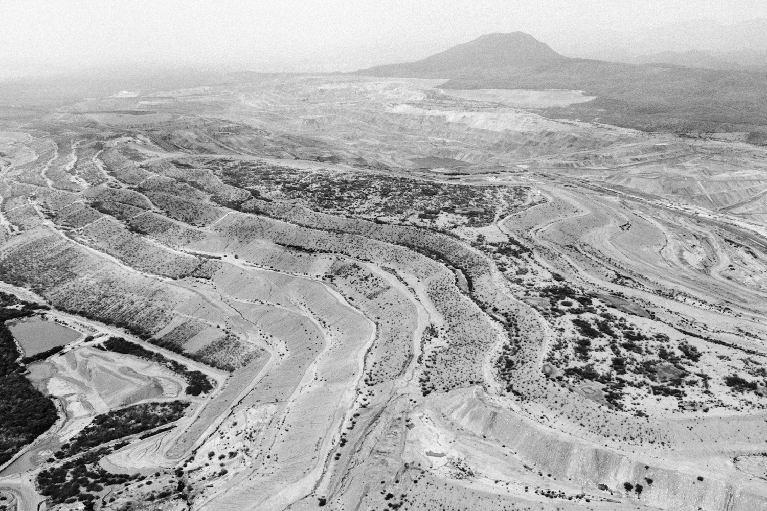 El Cerrejón: An expansive aerial view of El Cerrejón in La Guajira, Colombia, showing the immense scale of the open-pit coal mine. The deep, terraced industrial excavation cuts sharply into the earth, contrasting with the surrounding arid landscape of northern Colombia.. It is the largest open-pit mine in Latin America and one of the largest in the world, covering an area of roughly 690 square kilometers . The operation is completely integrated, featuring the mine itself, a dedicated 150 km (93-mile) private railway line, and its own private Caribbean export terminal called Puerto Bolívar. The mine is 100% owned and operated by Glencore PLC, a Swiss multinational commodity trading and mining giant.