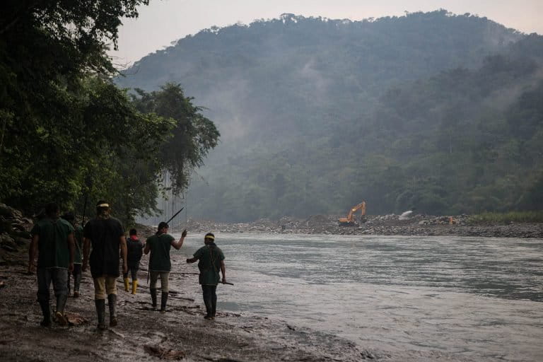 Miembros de la comunidad indígena Cofán de Sinangoe camina a orillas del río Aguarico, mientras al fondo se ve una retroexcavadora removiendo tierra. Foto: Jerónimo Zuñiga/Amazon Frontlines