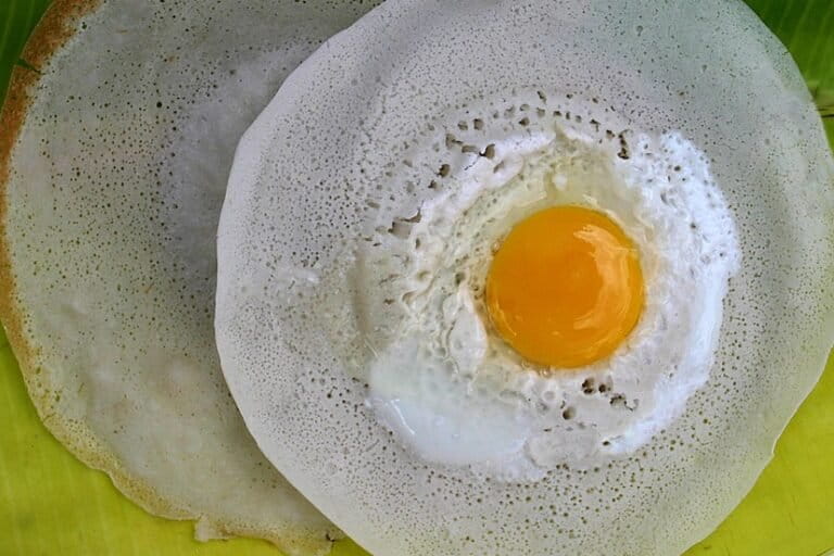 Image shows a fried egg on an appam on a palm leaf plate