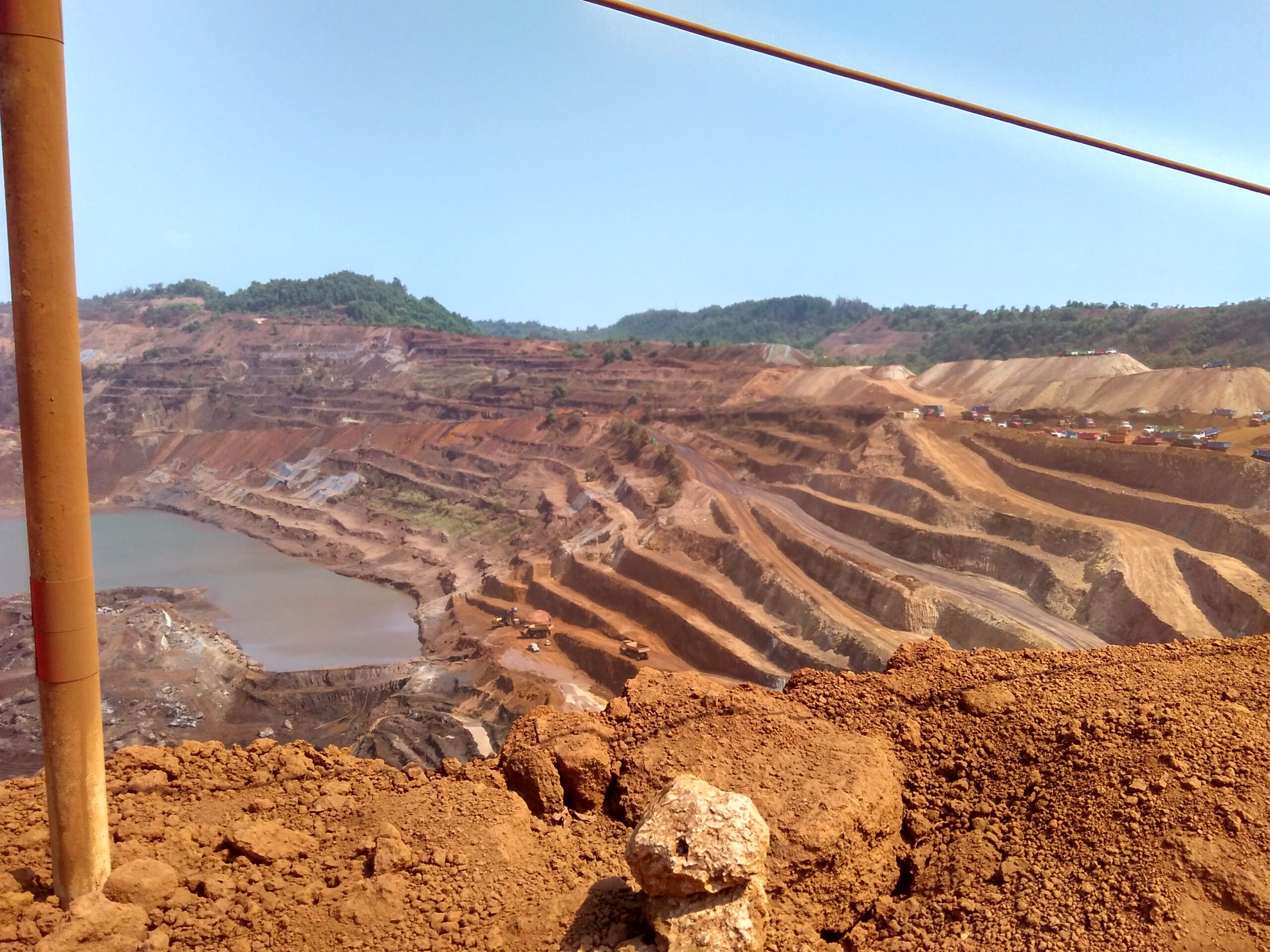 Image shows a deep mining pit surrounded by layers of dug-up soil and trees in the background
